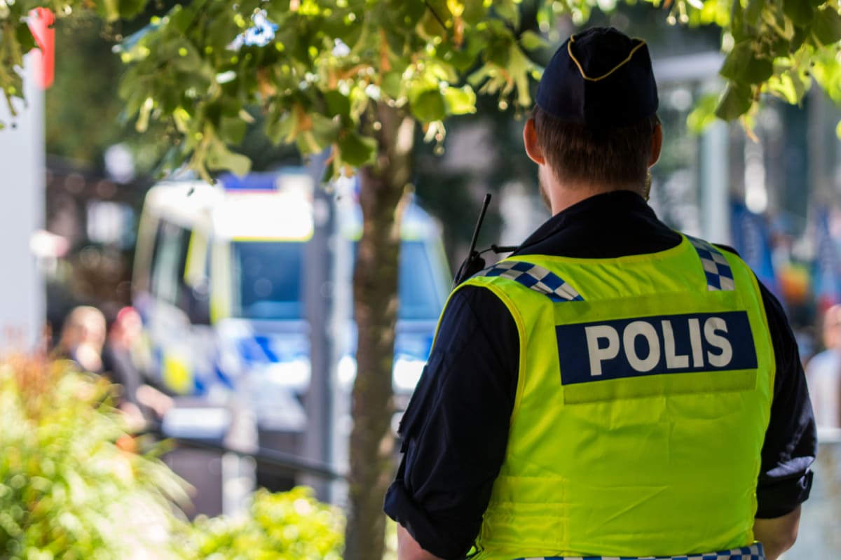 En bild tagen bakifrån på en person i polisuniform med en signalgul väst med texten "POLIS" i versaler. Personen bär en mörkblå uniformsmössa och en radio är fäst vid axeln. I bakgrunden syns suddiga gröna löv och ett fordon som liknar en ambulans el