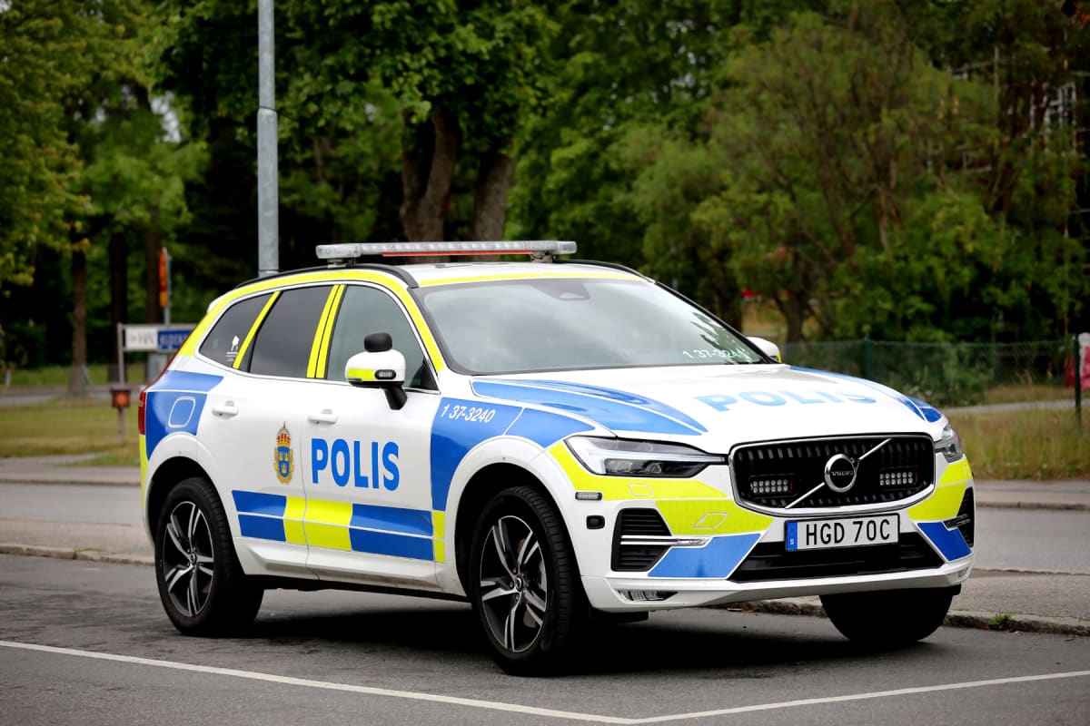 En svensk polisbil av märket Volvo står parkerad utomhus, delvis vänd mot betraktaren. Bilen är vit med blå och gula rutor och texten POLIS på sidan och motorhuven. I bakgrunden syns gröna träd och en stolpe.
