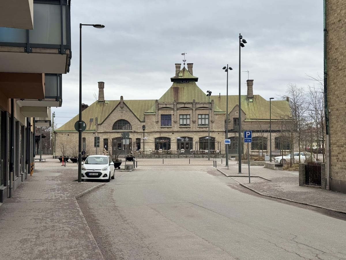 En ganska bred gata med en byggnad i bakgrunden. Byggnaden har ett grönt tak och verkar vara gjord av sten. Det är en skylt med P som betecknar parkering och bredvid den står ett träd. På gatan står också en vit bil. En annan byggnad syns på höger si