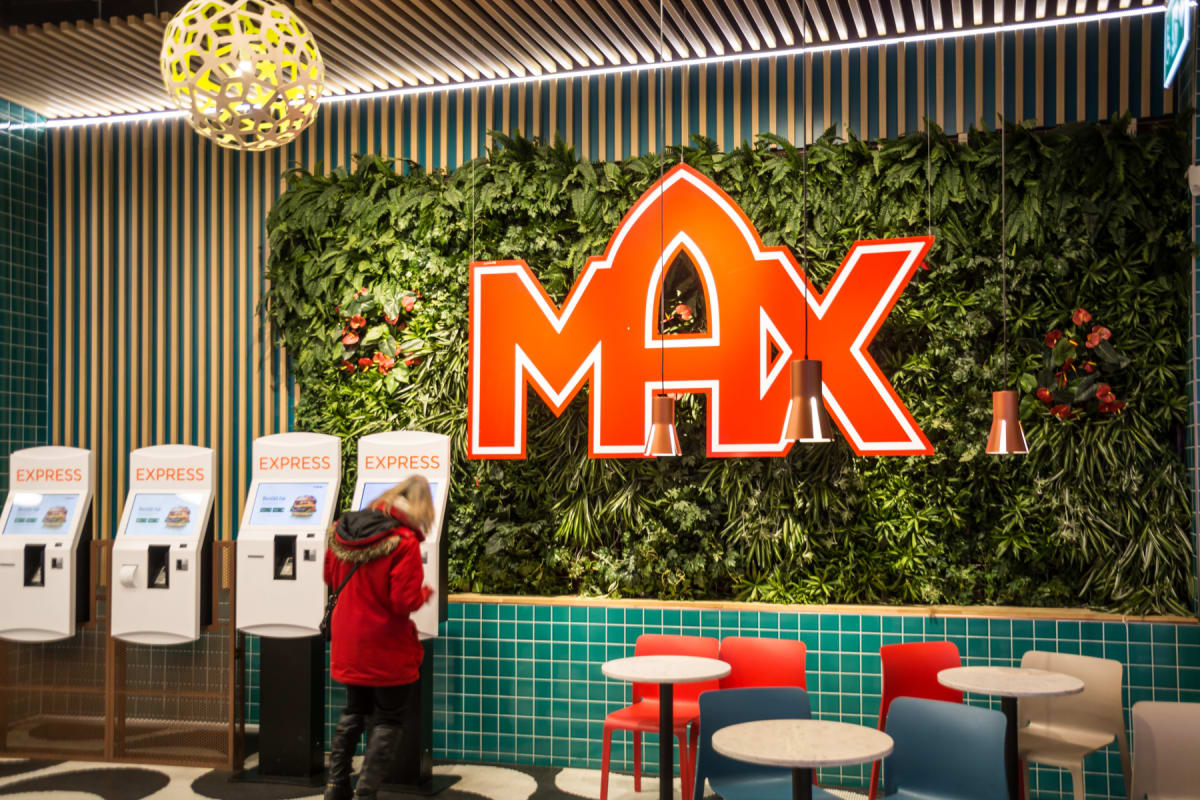 En bild inifrån en restaurang där en person i röd jacka beställer mat från en av fyra självbetjäningsmaskiner. En stor röd MAX-logotyp är monterad på en grön växtvägg, och flera bord med stolar syns i förgrunden.