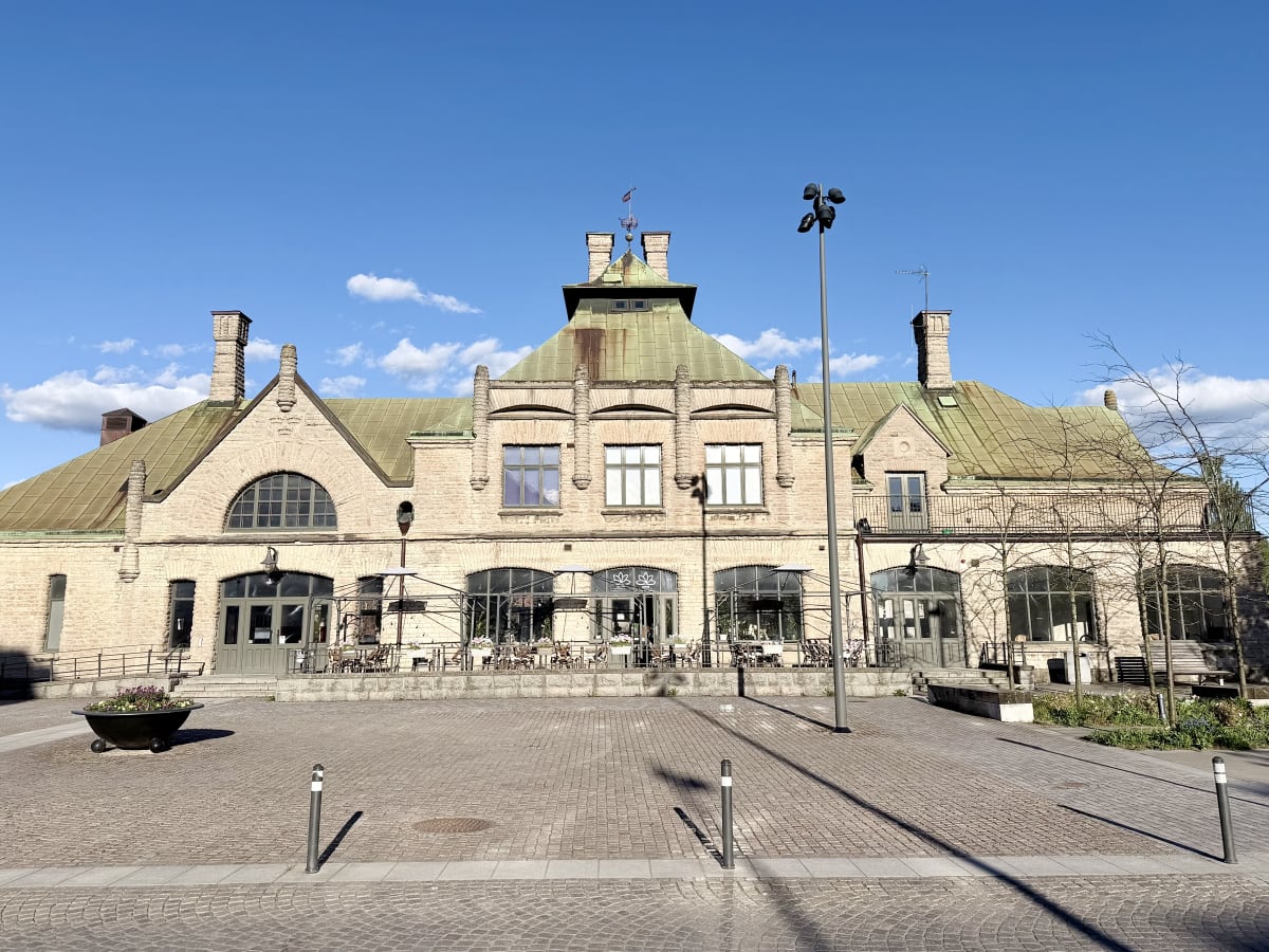 En stor, ljus tegelbyggnad med ett grönt plåttak under en klarblå himmel med några vita moln. Framför byggnaden finns ett öppet torg med kullersten. Byggnaden har flera fönster och dörrar samt en uteservering med bord och stolar. Det finns även stolp