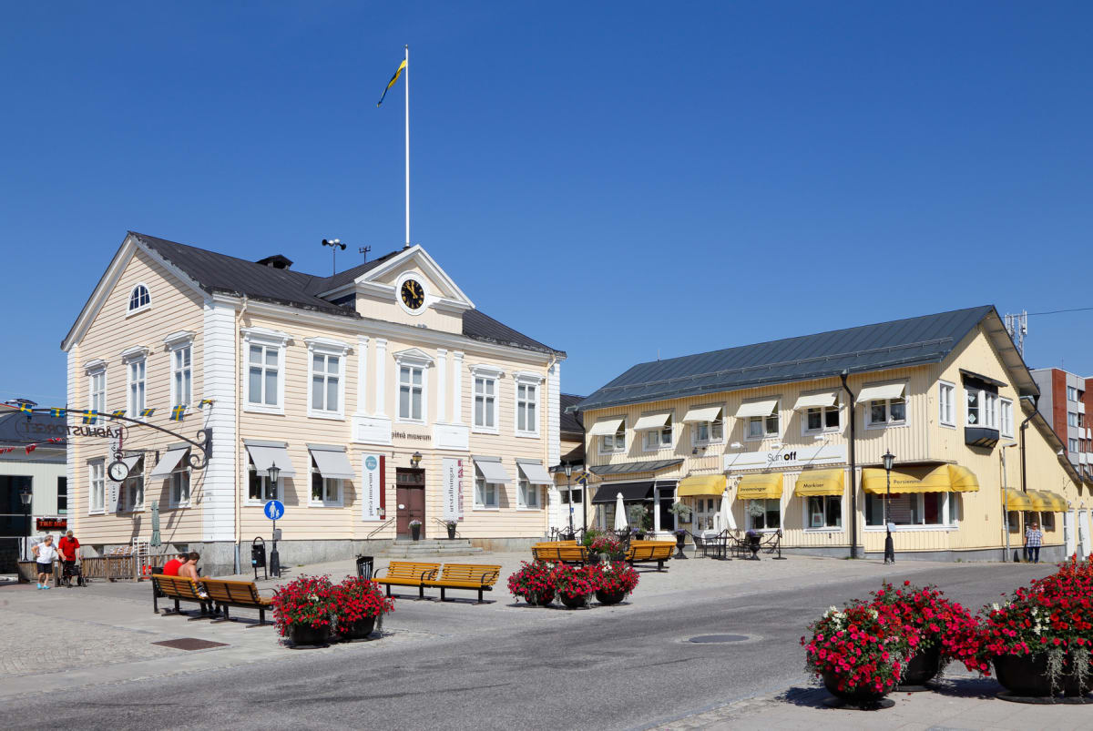 En central gata eller torg med en tidstypisk byggnad i ljusgul färg. Svenska flaggan syns i topp. Flera människor sitter på bänkar med blommor planterade framför. Byggnaden till höger är också ljus med gula markiser. Långa skuggor tyder på en stark s