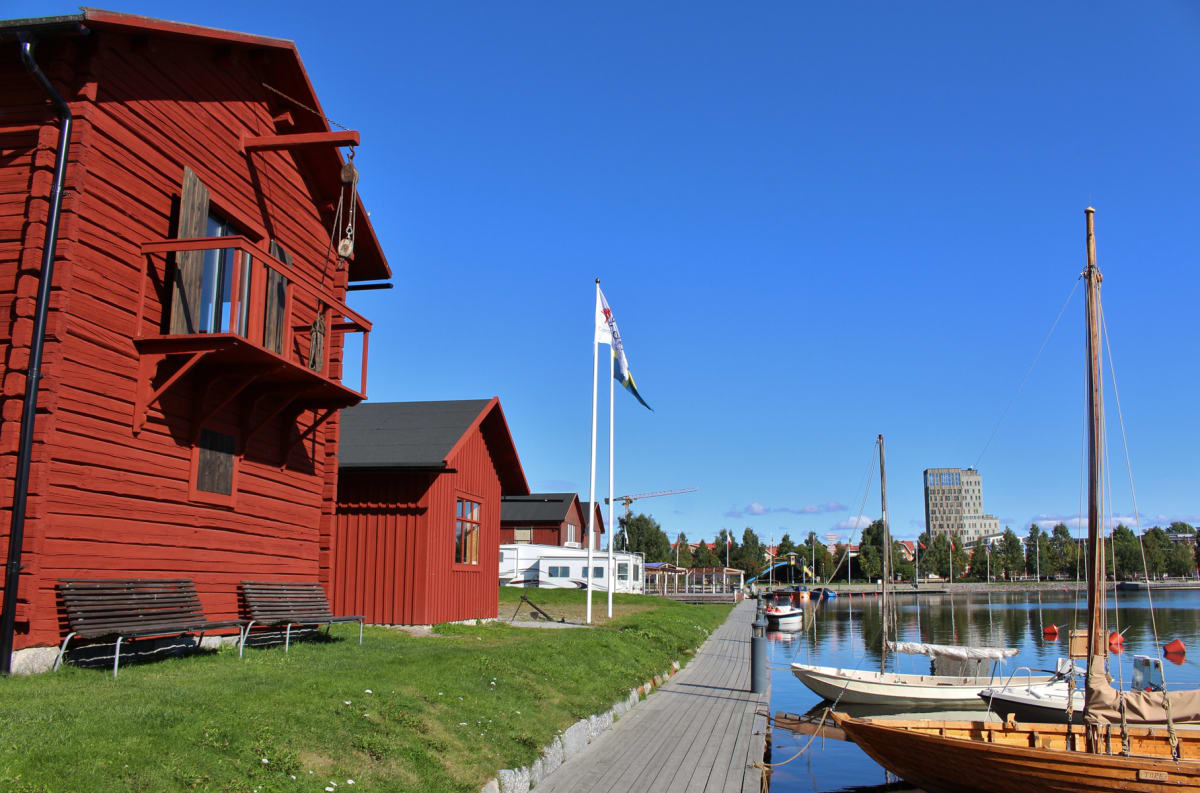 En bild som visar röda trähus med svarta tak vid en kaj med en träbrygga. Flera båtar ligger förtöjda i vattnet, och i bakgrunden syns skog samt en modern byggnad. En flaggstång med en flagga syns också.