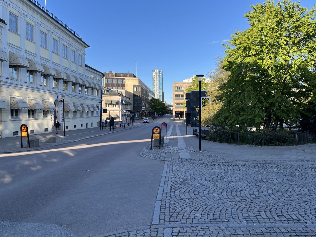 En gata i stadsmiljö med byggnader, träd och skyltar under en klarblå himmel. Till vänster syns en ljus byggnad med flera fönster och markiser. Längre fram på gatan står en hög byggnad och en person går på trottoaren till vänster.
