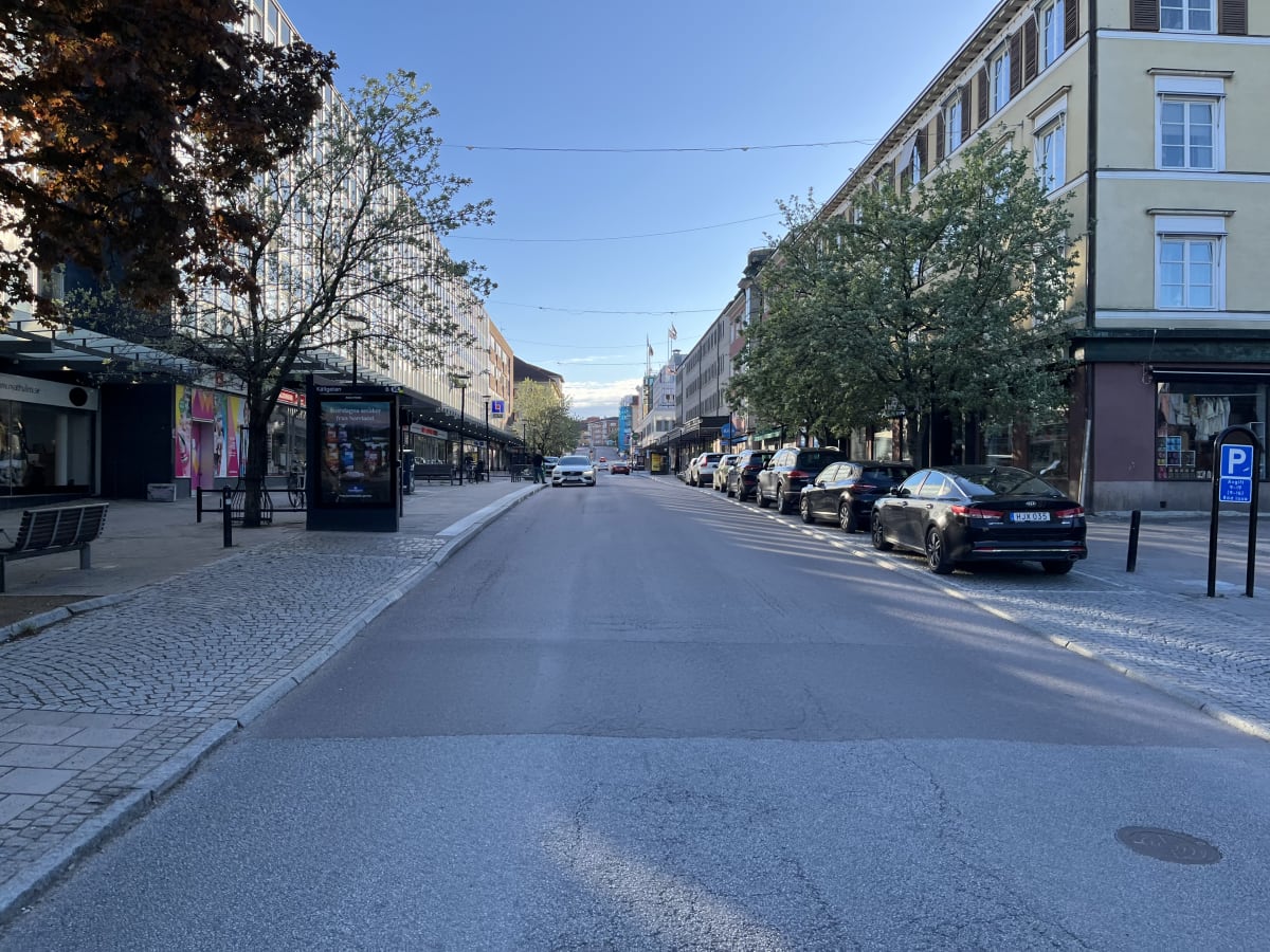 Bilden visar en gata i stadsmiljö med byggnader på båda sidor. På vänster sida finns träd med grönskande löv och butiksfasader. På höger sida står flera parkerade bilar längs en trottoar med stenläggning. På gatan syns en ensam bil nära centrum. Himl