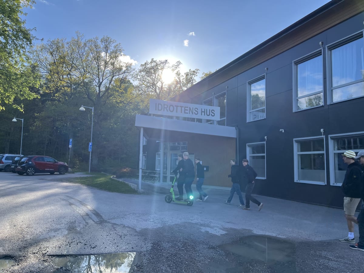 En utomhusbild tagen under dagtid föreställande entrén till en byggnad med skylten "IDROTTENS HUS". Flera personer syns framför byggnaden, varav en åker elsparkcykel. En röd bil är parkerad till vänster och träd syns i bakgrunden.