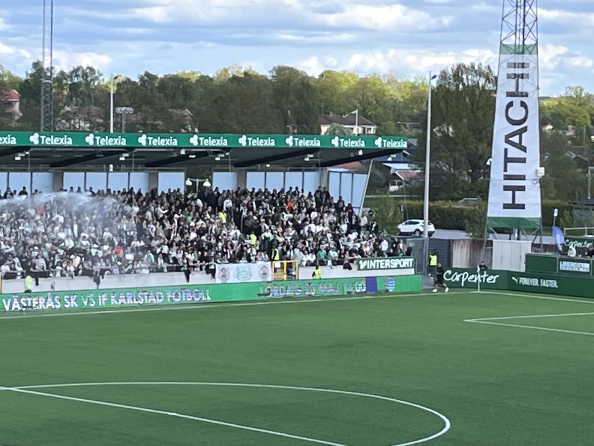 En utomhusbild föreställande en folkmassa på en arena där en fotbollsmatch spelas. Publiken sitter på läktare och nedanför dem syns en grön fotbollsplan med vita linjer. En skylt med "VÄSTERÅS SK VS IF KARLSTAD FOTBOLL" är synlig.