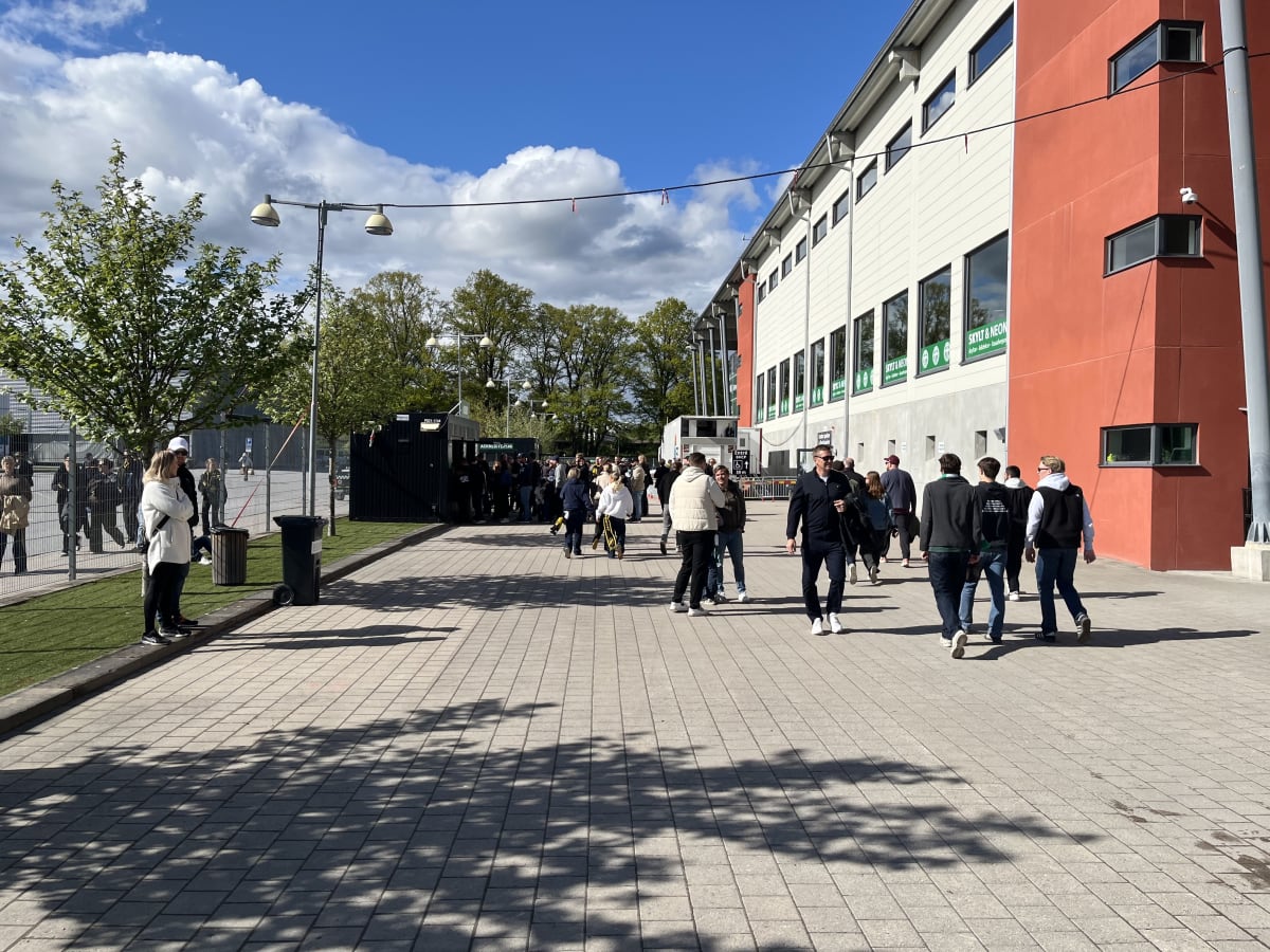 En utomhusbild som visar en grupp människor som går på en stenlagd gångväg bredvid en stor byggnad i ljust och rött. Människorna är klädda i fritidskläder och dagsljuset kastar skuggor från träd och byggnader. En rad av svarta containrar syns i bakgr