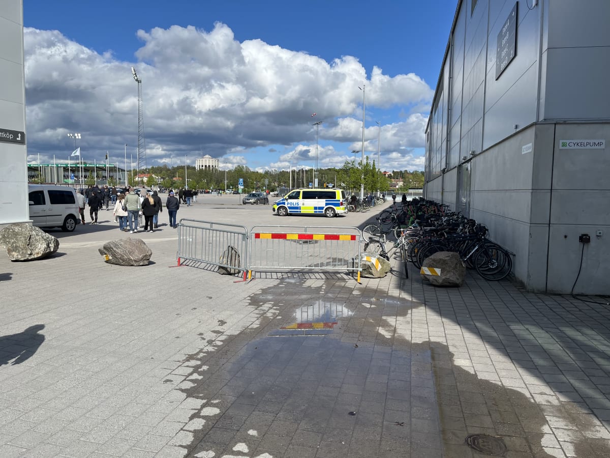 En utomhusbild som visar en stor folkmassa vid en byggnad. En polisbil, cykelställ och avspärrningar syns. På himlen syns både blå himmel och moln. Vid byggnaden står cyklar parkerade och en "CYKELPUMP"-skylt syns.