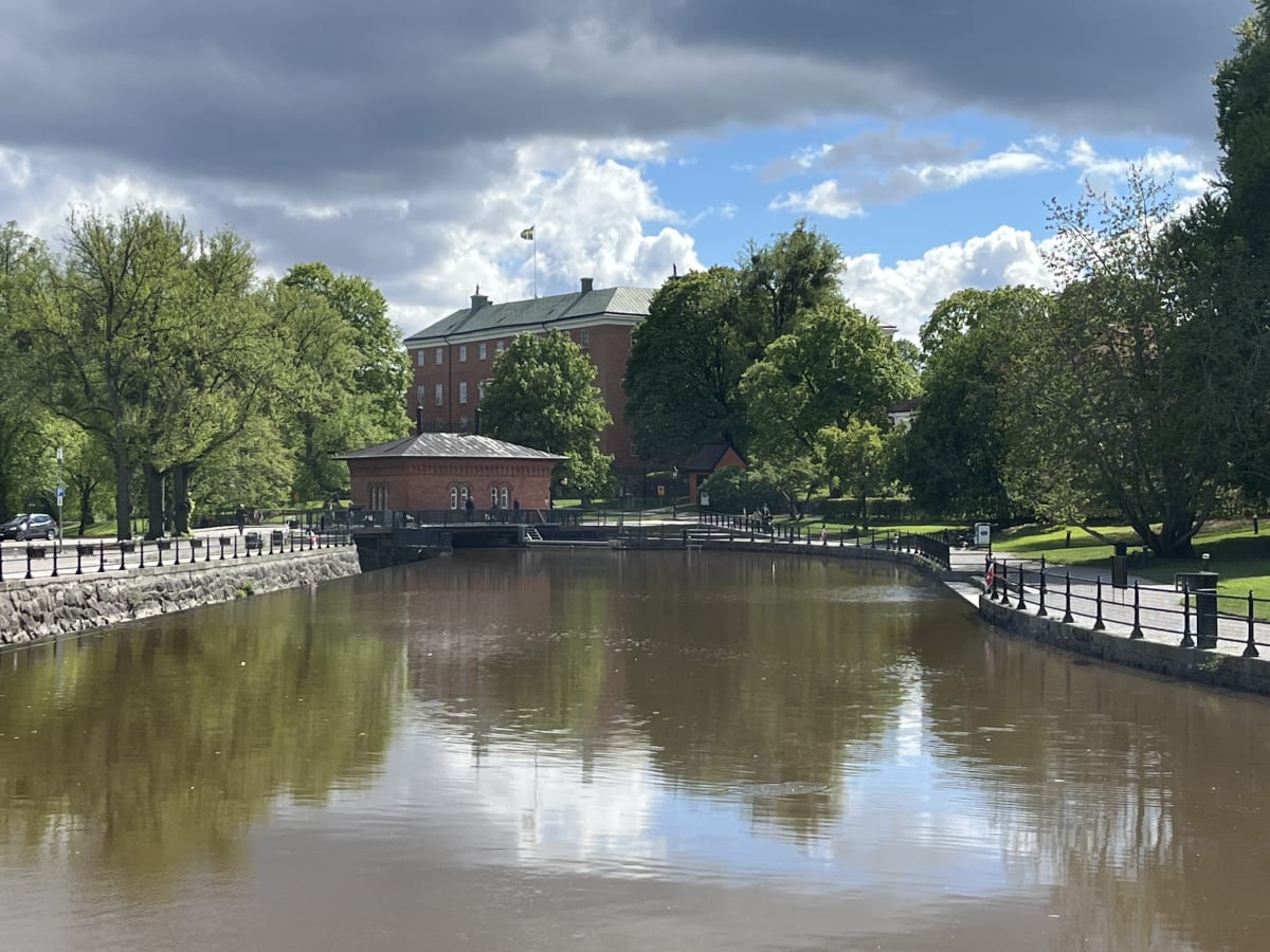 En bild en kanal med brunaktigt vatten omgiven av grönska och byggnader under en delvis molnig himmel. Två personer promenerar längs kanalen, och en stor byggnad med en svensk flagga syns i bakgrunden.