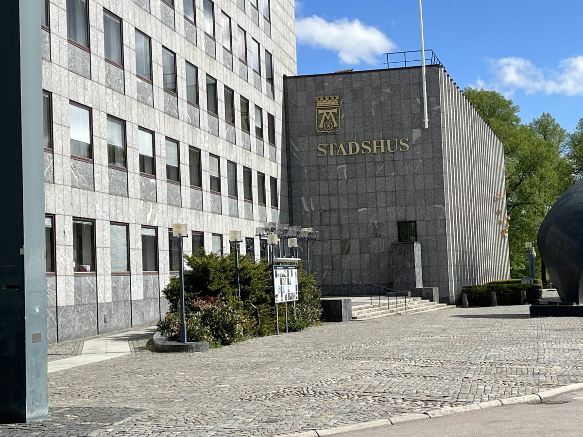 En ljus byggnad med många fönster till vänster, och en mörkare byggnad med texten "STADSHUS" och en symbol ovanför till höger. Träd med gröna löv syns bakom den mörkare byggnaden. Framför byggnaderna finns en yta med kullersten, buskar och en informa