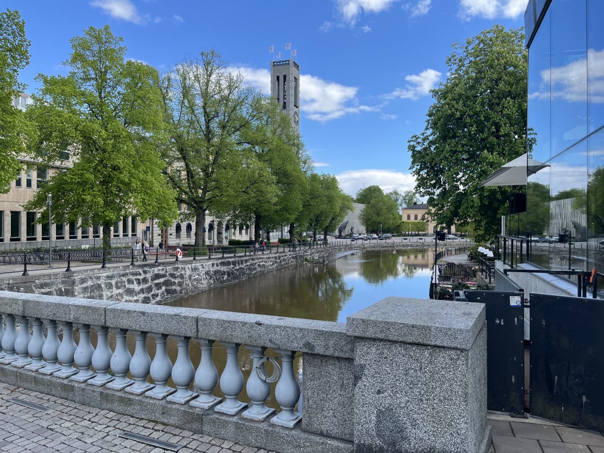 En bild tagen från en stenbro som vetter mot en kanal med byggnader och träd längs sidorna, inramad av en blå himmel med vita moln. I bakgrunden syns ett klocktorn och en modern byggnad i glas med speglingar.
