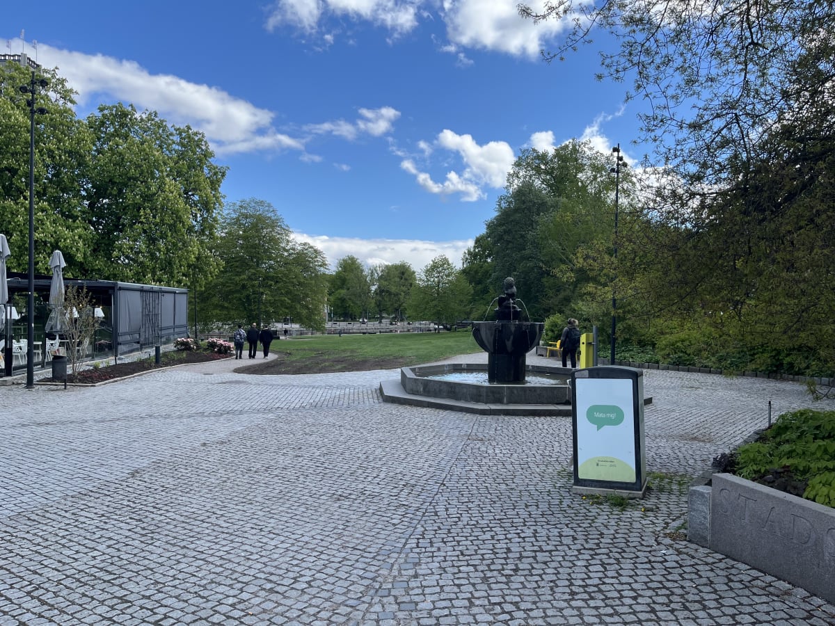 En utomhusbild som visar en kullerstensbelagd gångväg i en parkmiljö med gröna träd och en blåhimmel med vita moln. I mitten av bilden finns en fontän. Till höger finns en reklamskylt med texten "Mata mig!". Flera personer syns i bakgrunden.