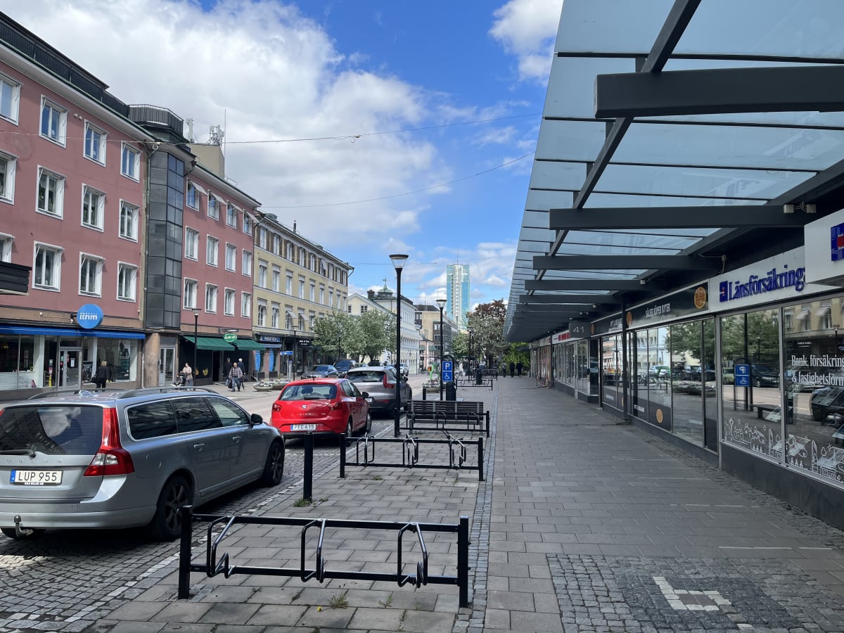 En gata med parkerade bilar, butiker och byggnader under en delvis molnig himmel. På vänster sida syns flerfamiljshus och butiker, medan höger sida har en modernare byggnad med en lång baldakin. Det finns även cykelställ och bänkar längs trottoaren.