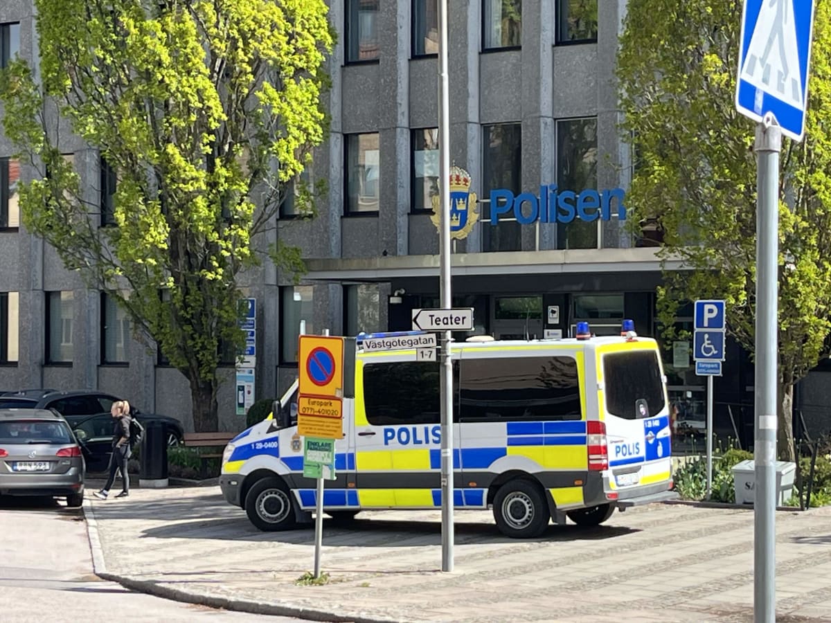 En polisfordon står parkerad nära en byggnad med texten "Polisen" och dess emblem. I förgrunden syns parkeringsskyltar och en person samt en bil i bakgrunden. Träd med gröna löv syns på båda sidor om byggnaden.