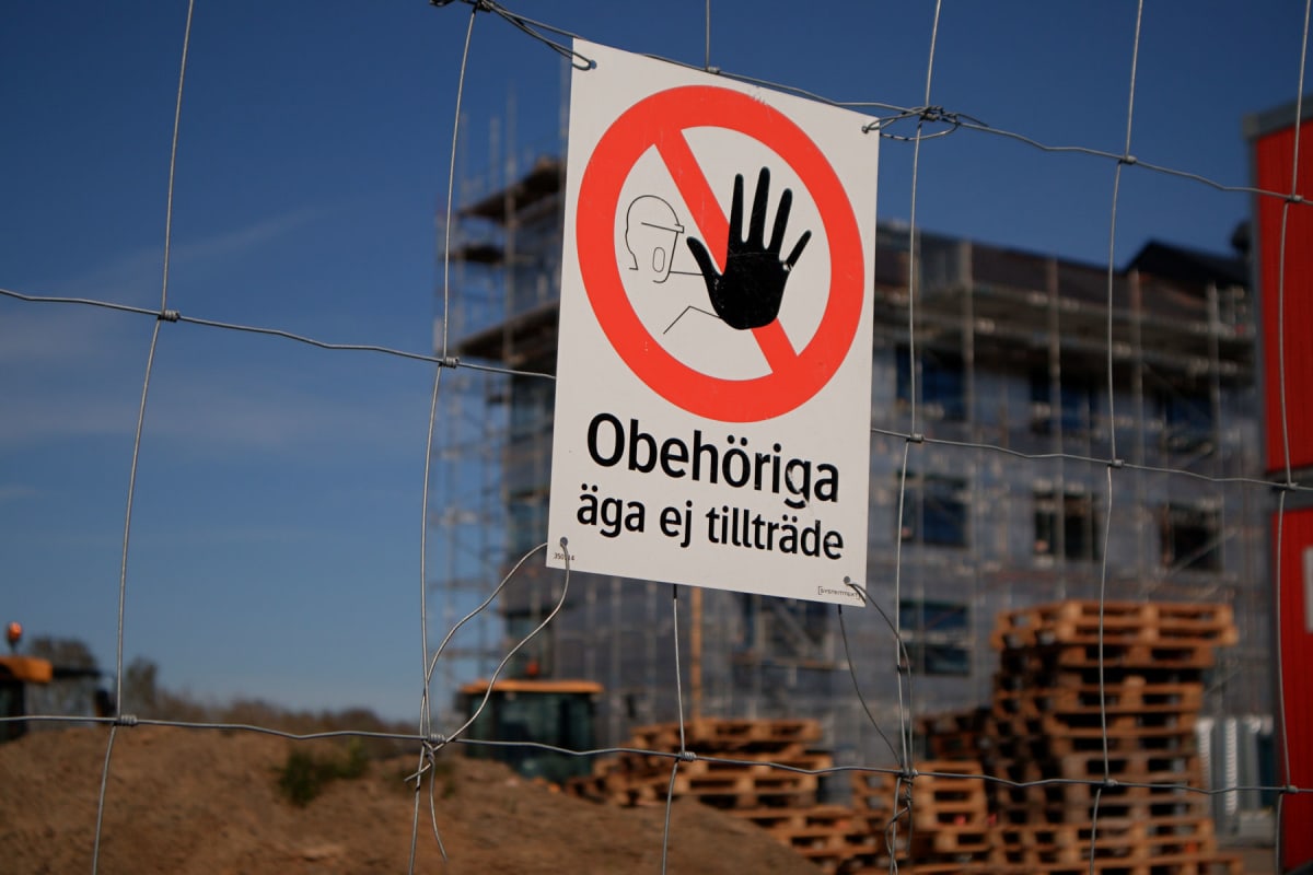 En bild som visar en skylt fäst vid ett stålstängsel. Skylten har en röd cirkel med ett svart streck diagonalt över en figur som signalerar stopp med handen som texten "Obehöriga äga ej tillträde". I bakgrunden syns en byggarbetsplats med ett oklart