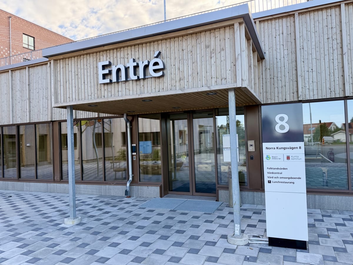 En modern byggnadsentré med träpanel och stora glaspartier under en skärmtak. Skylten "Entré" syns tydligt ovanför dörrarna. Till höger finns en stor informationsskylt med text om Folktandvården, Vårdcentral, Vård- och omsorgsboende samt Lunchrestaur