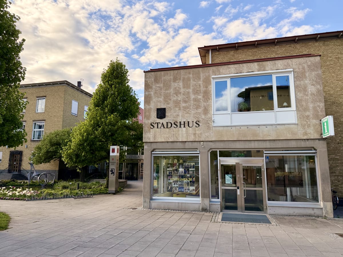 En bild av en byggnad med texten "STADSHUS" på framsidan. Ett turistinformationsmärke syns på höger sida. Framför byggnaden finns ett gångstråk och i bakgrunden syns fler byggnader och träd. Mellan byggnaderna syns en skylt som pekar mot "Stadshus en