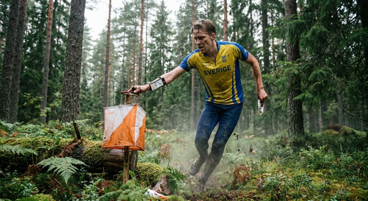 En man i sportkläder med texten "SVERIGE" på tröjan befinner sig i en skogsmiljö. Han håller i ett elektroniskt kontrollkort vid en orienteringsskärm. Mark och kläder är delvis smutsiga med jord och lera, vilket indikerar att han har rört sig i terrä