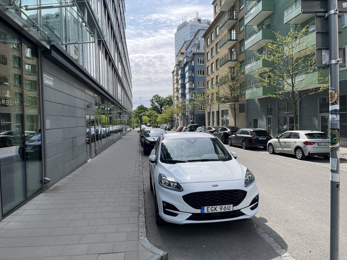 En gata inramad av byggnader med en rad parkerade bilar vid sidan. En vit personbil står i förgrunden till höger. På vänster sida av gatan syns en trottoar med en byggnad som har stora reflekterande fönster.