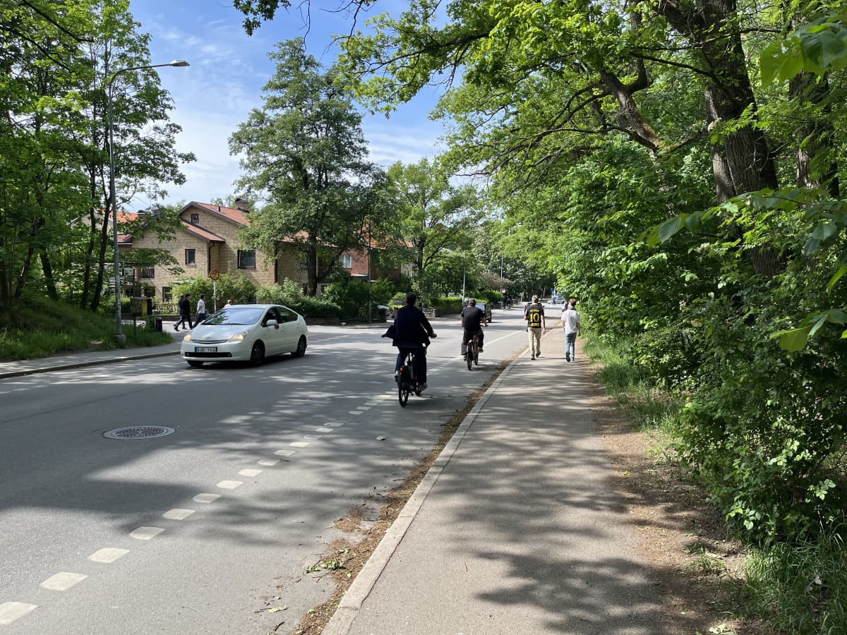 En gata med bilar och cyklister omgiven av träd och hus. En vit bil kör på vägen. Flera personer syns till fots och på cyklar. Långa skuggor faller över vägen och trottoaren, vilket indikerar soligt väder.