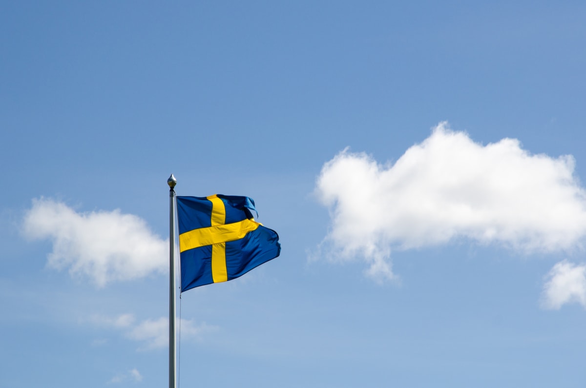Den svenska flaggan vajar på en flaggstång under en delvis molnig himmel. Flaggan är blå med ett gult kors och ses mot en klarblå himmel med några vita moln i bakgrunden. Flaggstången har en silverfärgad knopp högst upp.