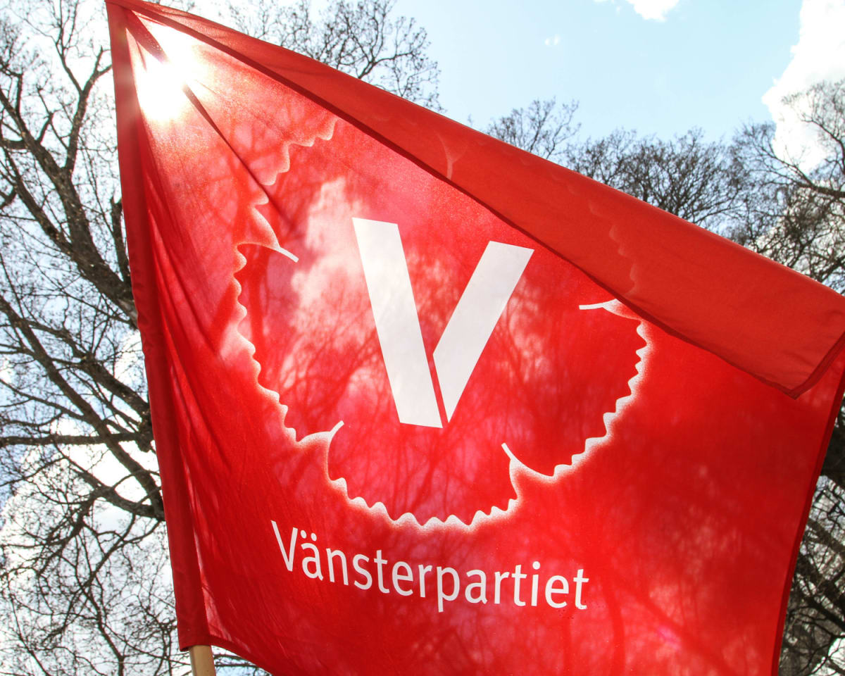 En röd flagga med texten "Vänsterpartiet" nedtill och en vit stiliserad V-logotyp mot en blå himmel med några moln. Solen reflekteras i flaggans övre vänstra del. Trädgrenar utan löv syns i bakgrunden.