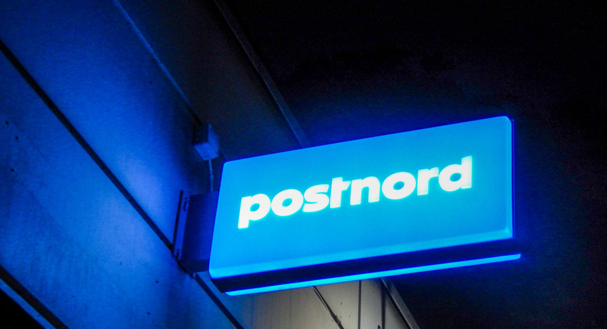 En upplyst skylt med texten "postnord" i vitt på en ljusblå bakgrund. Skylten är fäst vid en byggnadsfasad med horisontella paneler och detaljer i mörkblå nyanser. Det är mörkt runtomkring skylten, vilket indikerar att bilden är tagen nattetid eller
