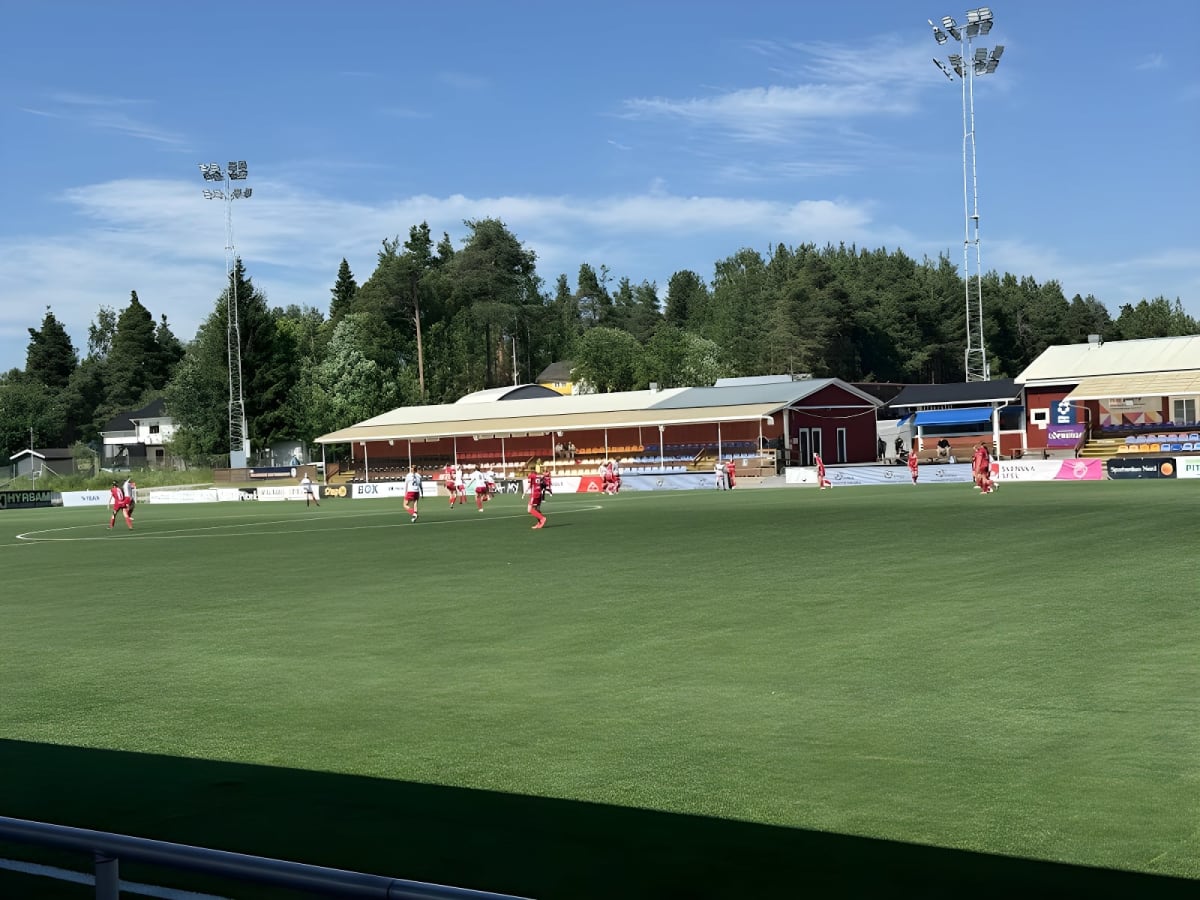En utomhusbild tagen under dagtid på en fotbollsplan med konstgräs, där spelare i röda och vita tröjor befinner sig på planen. I bakgrunden syns skogsbevuxna backar, byggnader, en läktare, och flera höga belysningsstolpar.