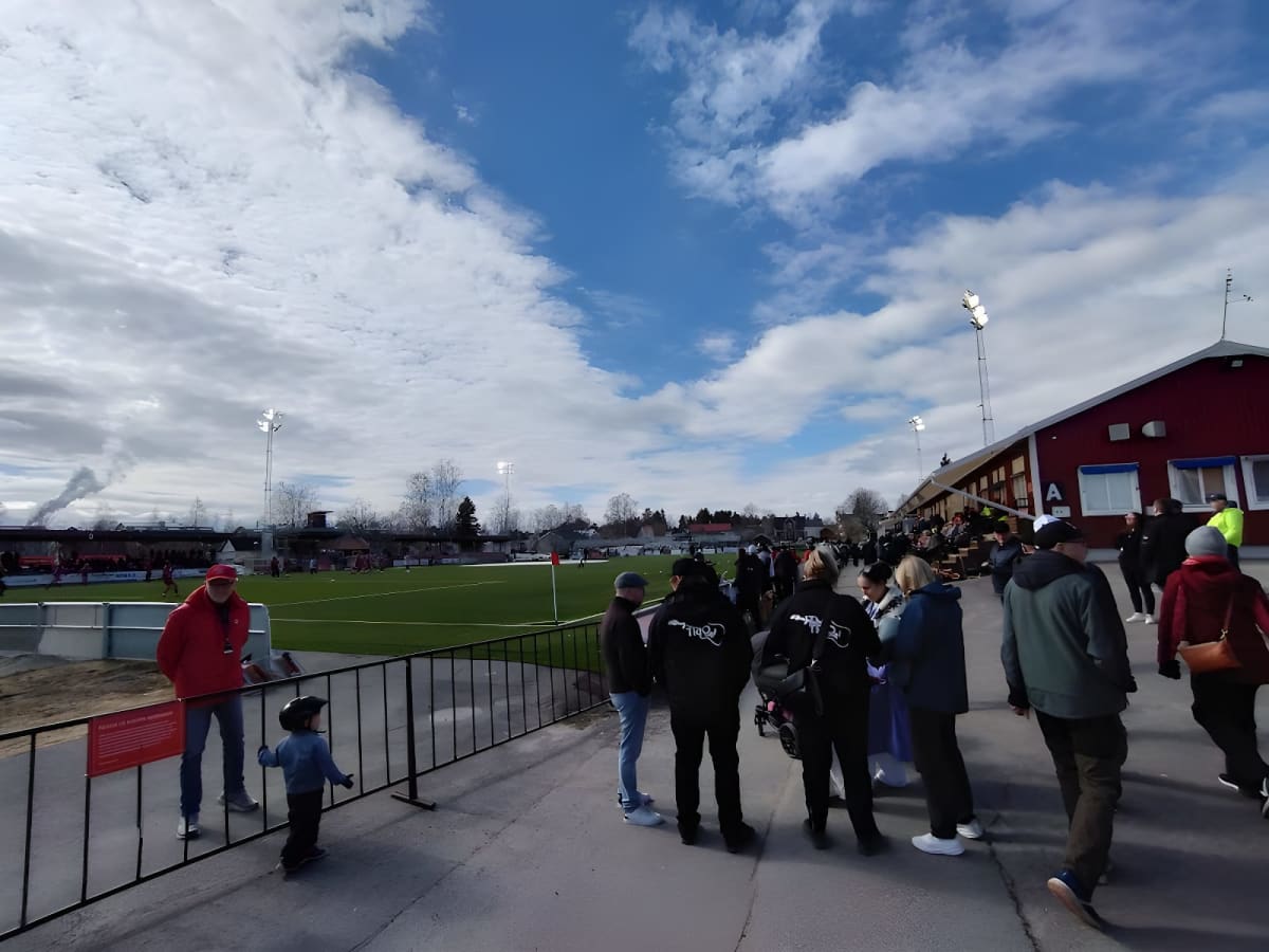 En utomhusbild som visar en grupp människor på en gångväg vid en fotbollsplan under dagtid. Bakom människorna syns en röd byggnad, strålkastare och en grön fotbollsplan. Himlen är blå med vita moln och rök syns i bakgrunden.