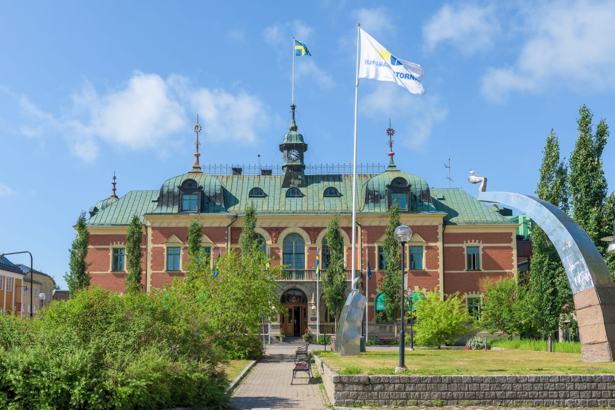 En stor, röd tegelbyggnad med grönbrunt koppartak dominerar bilden under en klarblå himmel. Framför byggnaden finns en gångväg med parkbänkar, gräsytor, träd och buskar. Två flaggstänger reser sig, en med svensk flagga och en med vit flagga med texte