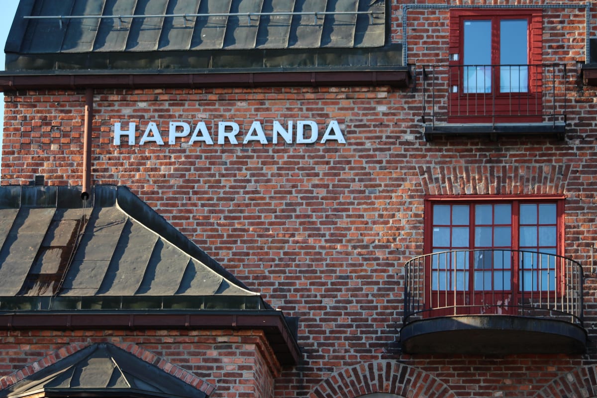 En fasad av rött tegel med vita bokstäver som bildar 'HAPARANDA' i mitten. På fasaden finns också en takränna, två rödfärgade fönster och en balkong med svart räcke. Taket är grått och syns i bildens övre och nedre delar.