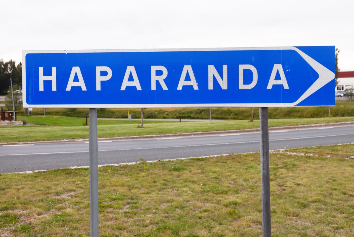 En blå skylt med vit text "HAPARANDA" och en vit pil som pekar åt höger, står på två grå metallstolpar. Bakom skylten syns en asfalterad väg med vita väglinjer och gräsmarker.