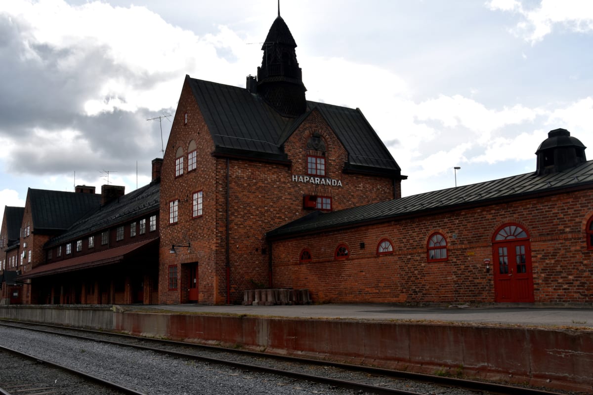 En stor, mörk byggnad av rött tegel med texten "HAPARANDA" syns på fasaden. Byggnaden har flera fönster med röda fönsterramar och svarta tak. I förgrunden syns tågspår med grus runtomkring och en perrong av betong. Himlen är delvis molnig.