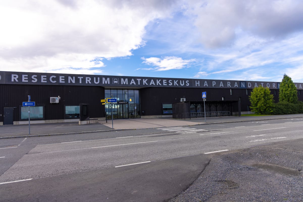 En exteriörbild av en svart byggnad med texten "RESECENTRUM MATKAKESKUS HAPARANDA" i stora vita bokstäver på fasaden. Framför byggnaden finns en asfalterad väg med vita vägmarkeringar och en trottoar. Blåa skyltar och grönska syns också.