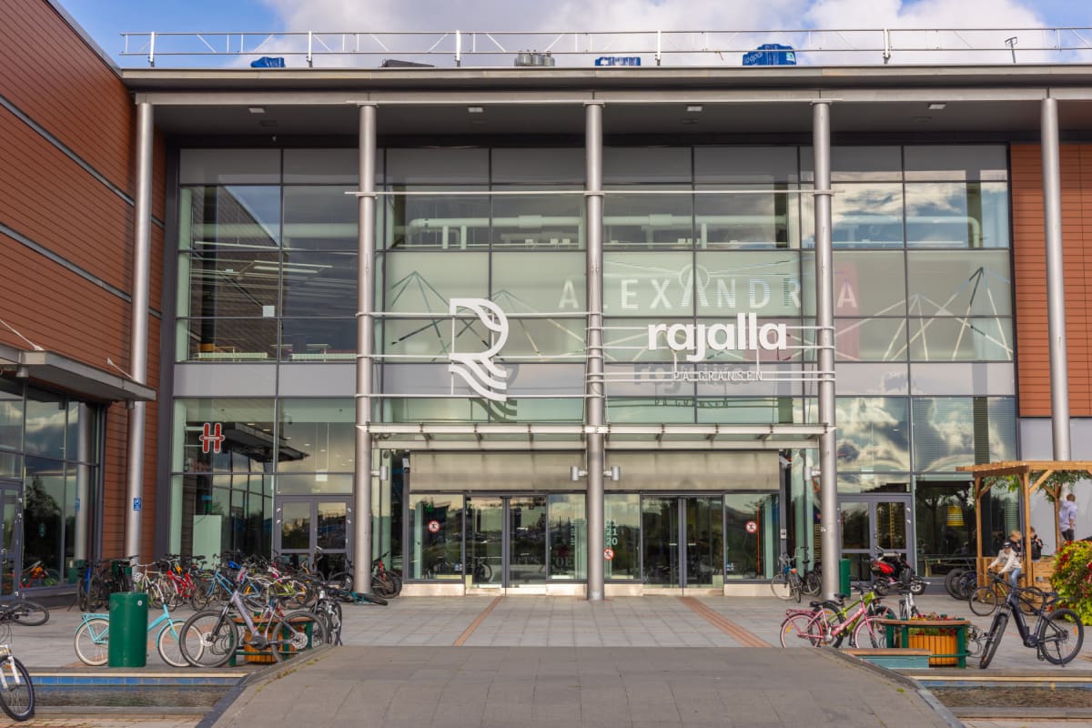 En modern byggnad med en fasad av glas och bruna paneler syns under en molnig himmel. Framför byggnaden finns cyklar parkerade. Övre delen av byggnadens glasfasad har vit text som lyder "ALEXANDRA rajalla PÅ GRÄNSEN". Flera personer syns vid en trädk