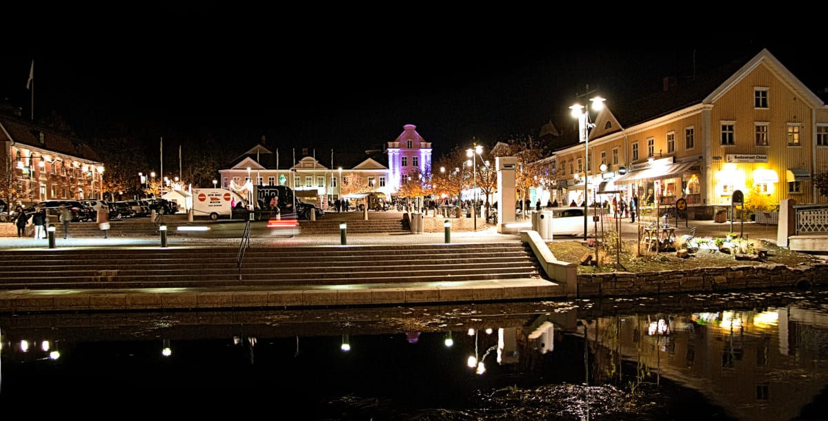 En nattbild visar en upplyst stadsmiljö med byggnader, en kanal eller å, trappor och människor. Gatljus och fasadbelysning reflekteras i vattnet. Ett släp med texten "NYR MIG!" och ett telefonnummer kan ses bakom bilar i vänster del av bilden.