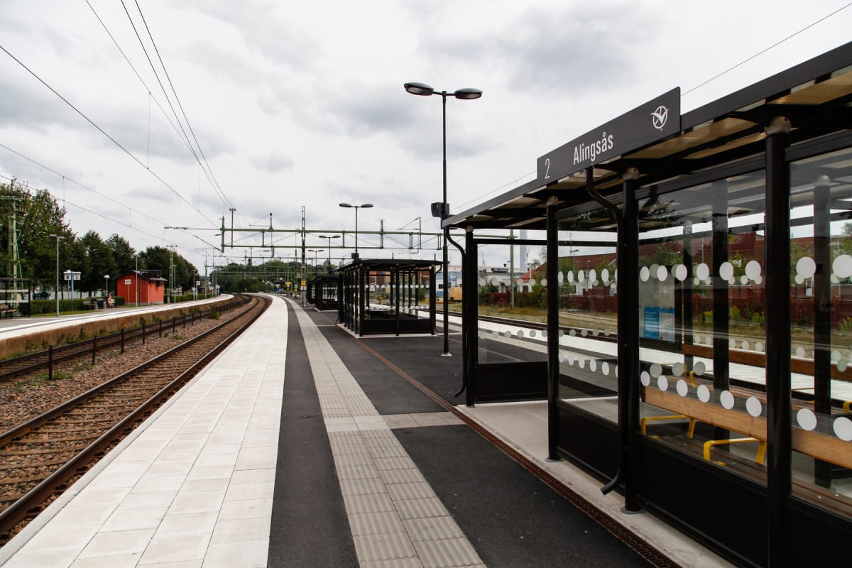 En bild av en järnvägsstation utomhus, tagen under dagtid. Två järnvägsspår syns, där det vänstra spåret är mest framträdande. Stationen har perronger med tak och väntkurer med bänkar. En skylt med texten "Alingsås" och siffran "2" är synlig. Ett bos