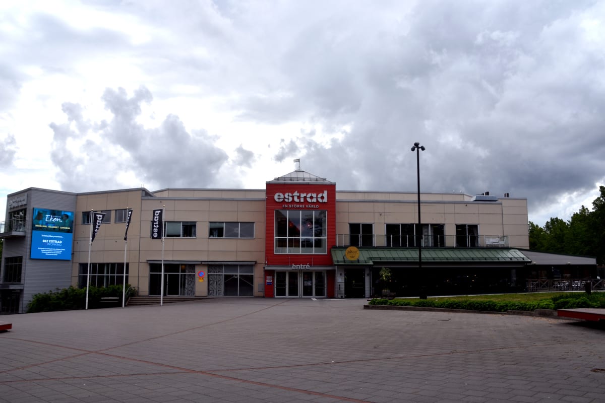 En stor, ljusbeige byggnad med en framträdande röd sektion i mitten, som har texten 'estrad' och 'EN STÖRRE VÄRLD' tryckt på sig. Byggnaden har många fönster. Framför byggnaden finns ett stort torg med gråa stenplattor och röda markeringar. Ett par f