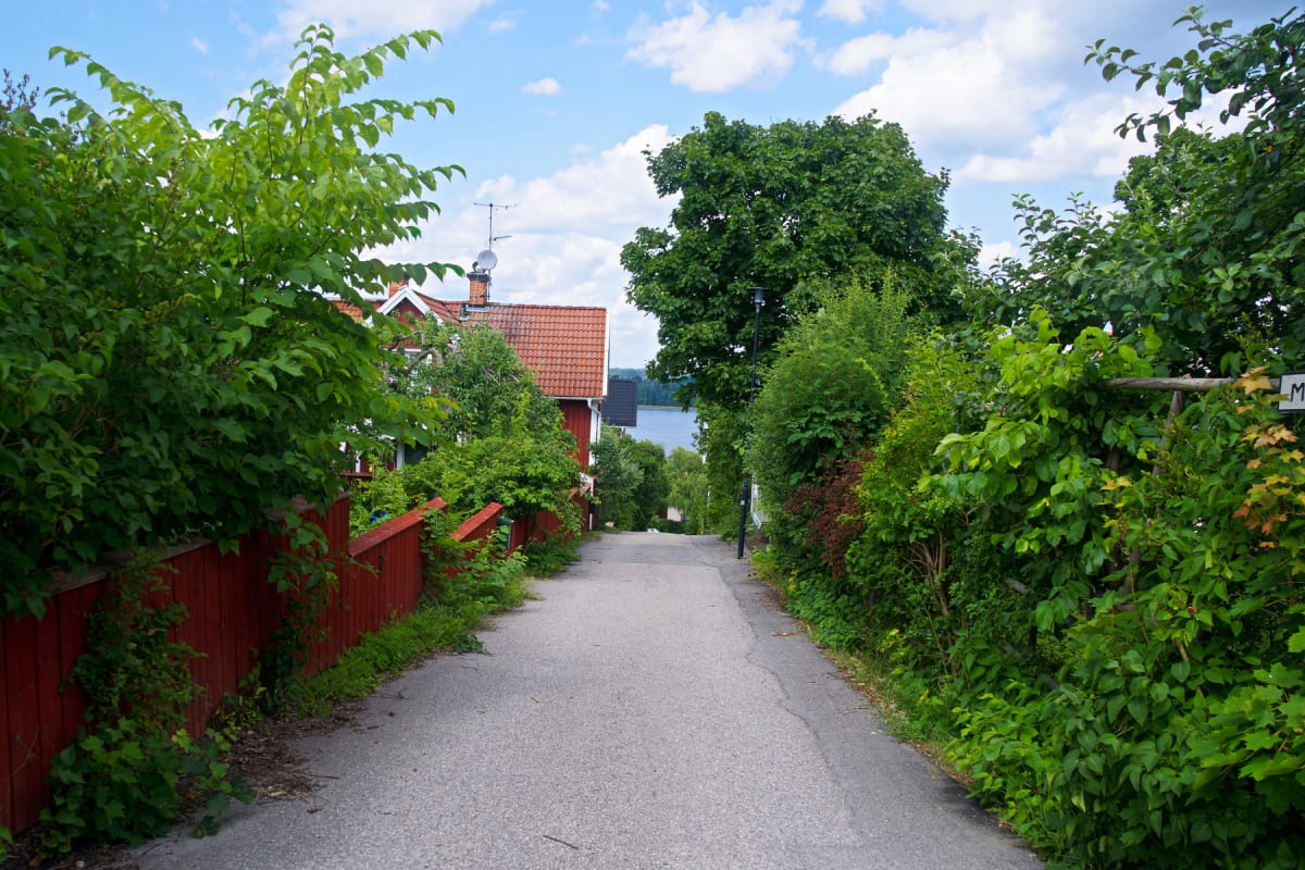 En nedåtgående asfalterad väg kantad av buskar och träd, som leder mot en sjö. På vänster sida av vägen syns ett rött trästaket och ett rött hus med tegeltak. Himlen är blå med vita moln.