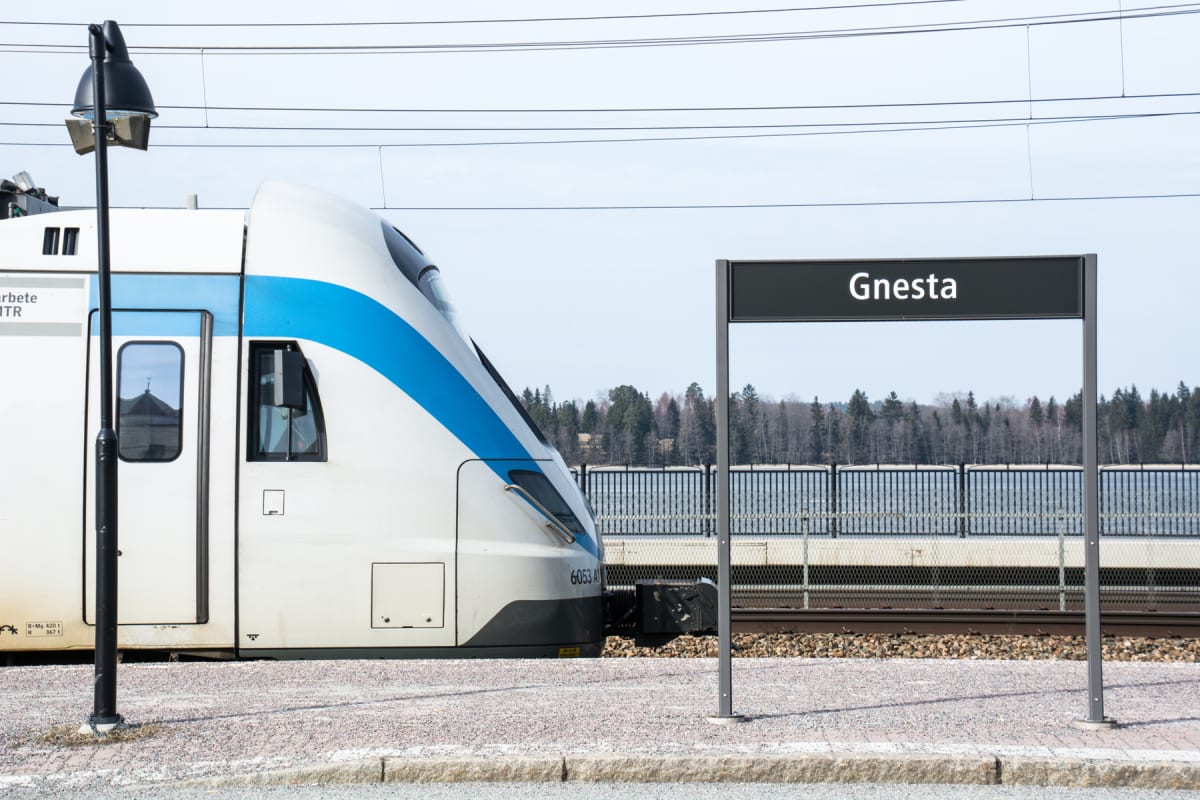 En vit och blå färgad pendeltågsvagn står vid en perrong. I förgrunden syns en svart skylt med texten "Gnesta". Bakom tåget och skylten sträcker sig ett staket, träd och en vattenyta. En del av tågets registreringsnummer "6053 A1" syns.