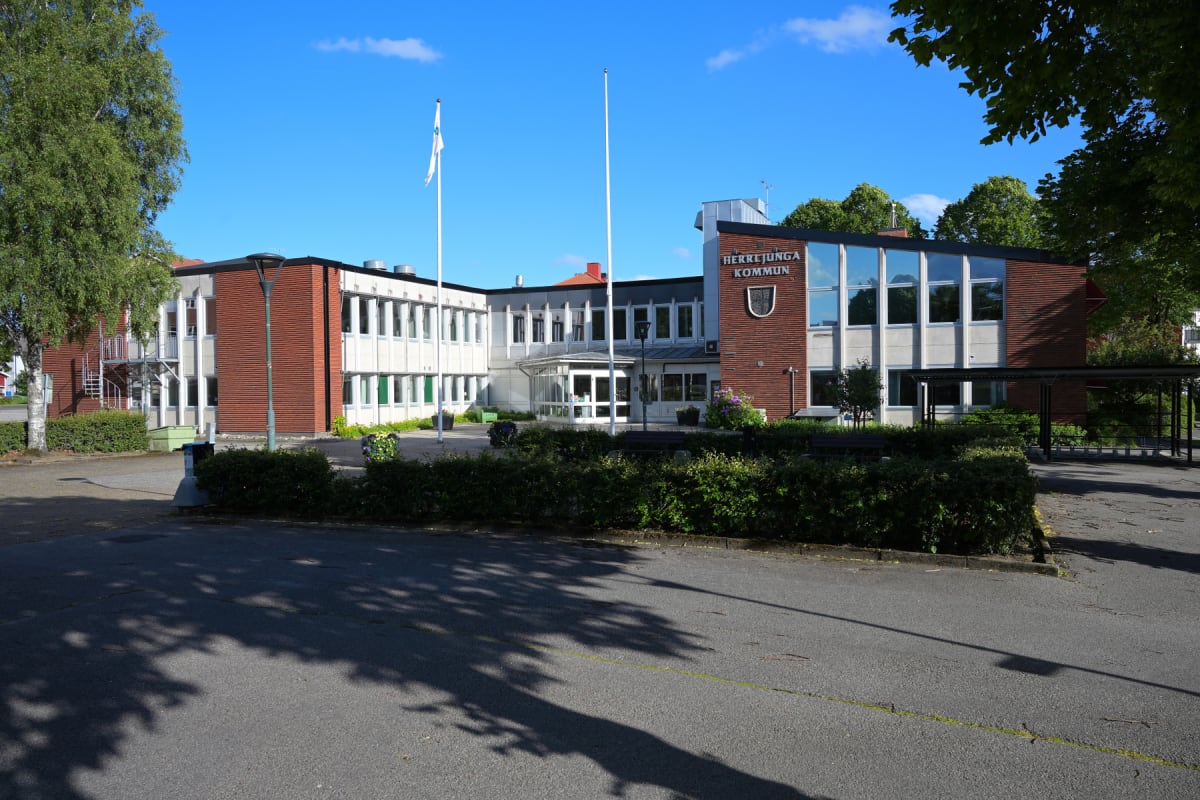 En stor, L-formad byggnad med tegelfasader och stora fönster, en del av fönstren är vita och omgivna av vita ramar, medan andra är stora, reflekterande fönster. Framför byggnaden finns ett asfalterat område, gröna buskar och träd. Två flaggstänger me