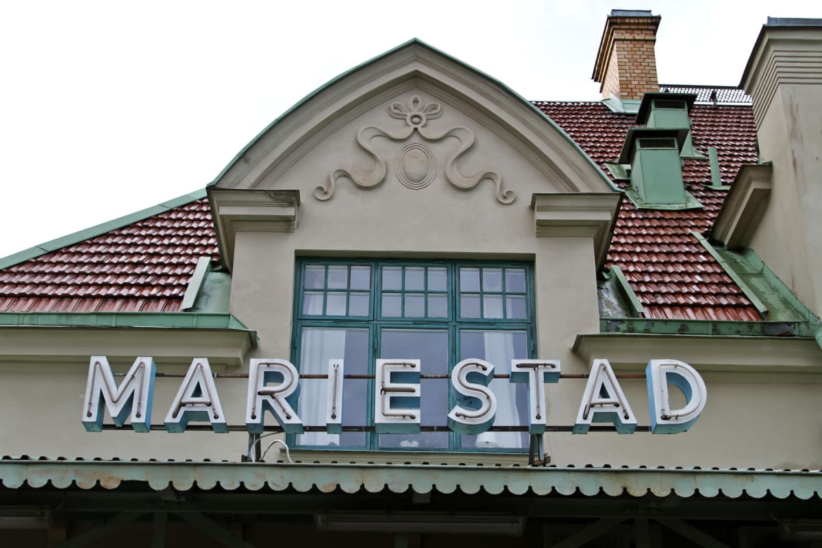 En fasad på en byggnad med ett stort fönster med spröjs. Ovanför fönstret finns dekorativ puts och ett tak med röda tegelpannor samt ett flertal grönmålade takdetaljer och en skorsten i tegel. Nedanför fönstret sitter bokstäverna 'MARIESTAD' monterad