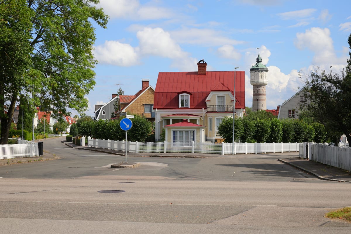 En gata framtill med hus och staket längs sidorna, samt träd med gröna löv. I bakgrunden syns fler hus med röda tak och ett högt vattentorn av tegel med en grön kupol. Himlen är blå med vita moln och det är ljust och soligt.