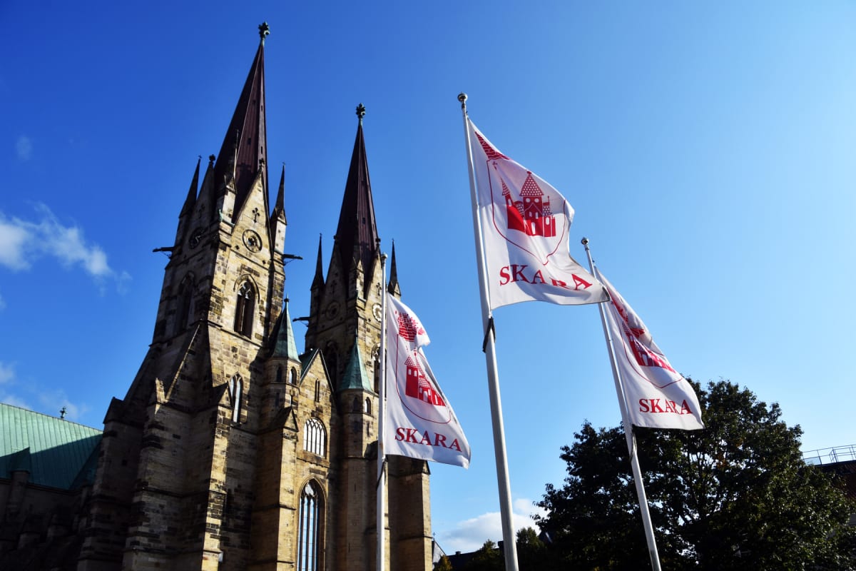 En stor, mörk kyrkobyggnad med höga spiror, omgiven av träd, står mot en klarblå himmel. Tre flaggor med texten "SKARA" och en röd symbol är placerade framför kyrkan, sedda nedifrån.