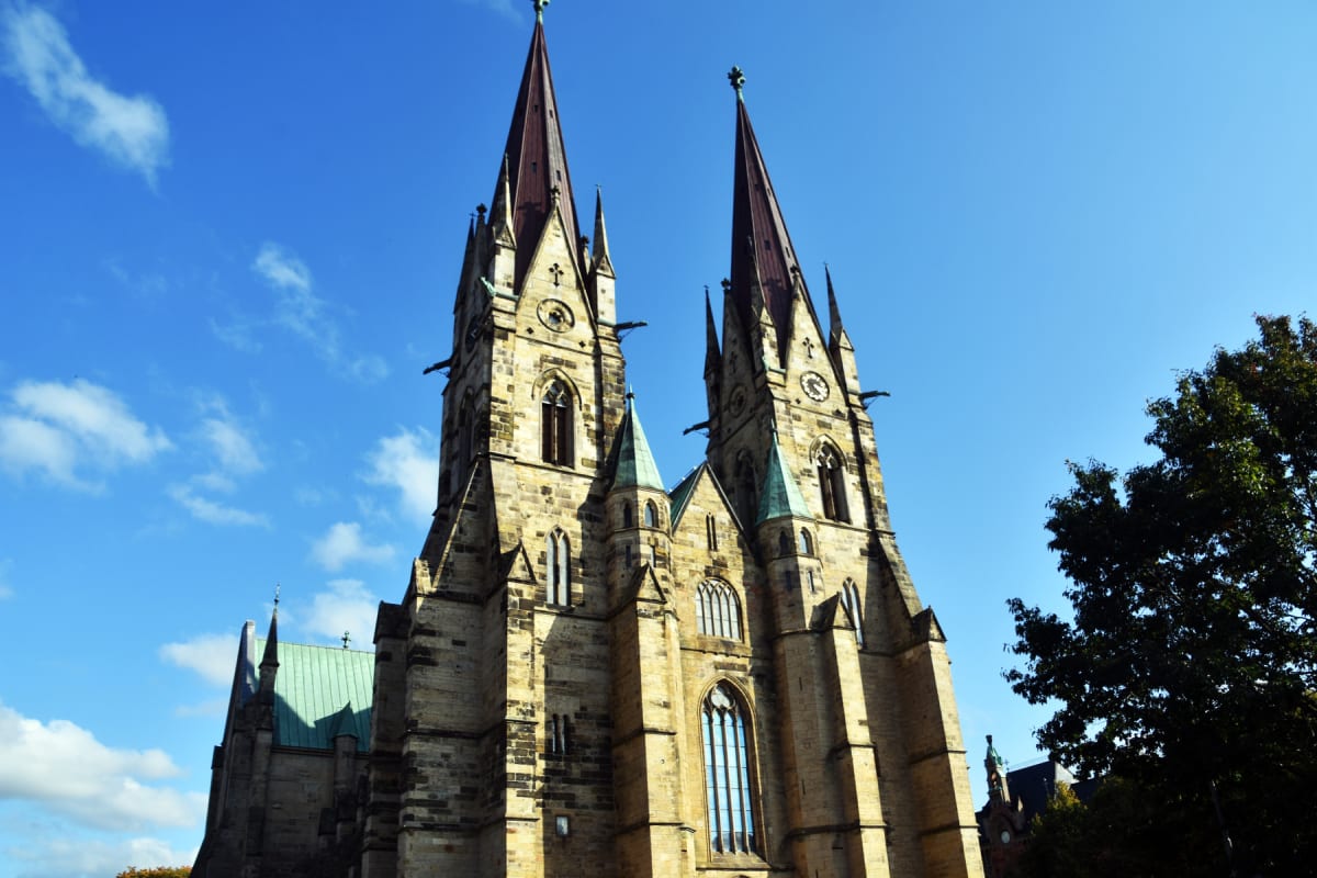 En stor, gotisk kyrkobyggnad med två höga spiror, flera fönster och klockor. Kyrkan är byggd i ljus sten och har gröna takdetaljer. En del träd syns till höger om byggnaden, mot en klarblå himmel med några vita moln.