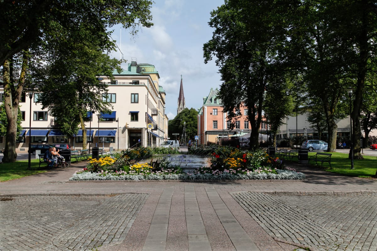 En central bild av en torgplats med en blommande fontän i förgrunden. Runt fontänen finns gräsmattor, träd och bänkar. På vardera sida syns byggnader, varav en större kyrkspira syns i bakgrunden. Två personer sitter på en bänk till vänster och en bil