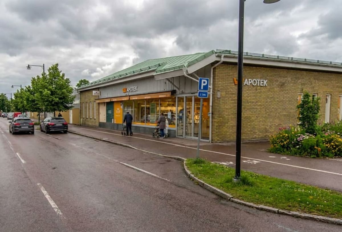 En gata med parkerade bilar leder fram till en byggnad med texten "APOTEK" på fasaden. Två personer använder rullatorer framför byggnaden. Ett P-skylt med tillägg "2 tim" står utanför entrén. Molnig himmel och grönska syns i bilden.