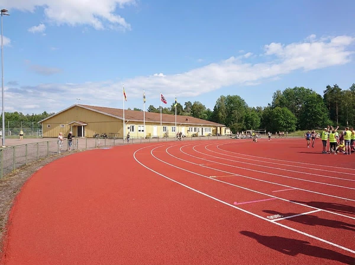 En utomhusbild som visar en röd löparbana med vita linjer, del av en idrottsplats. I bakgrunden syns en gul byggnad med flera flaggstänger framför, samt lövträd, en skog, och en blå himmel med vita moln.