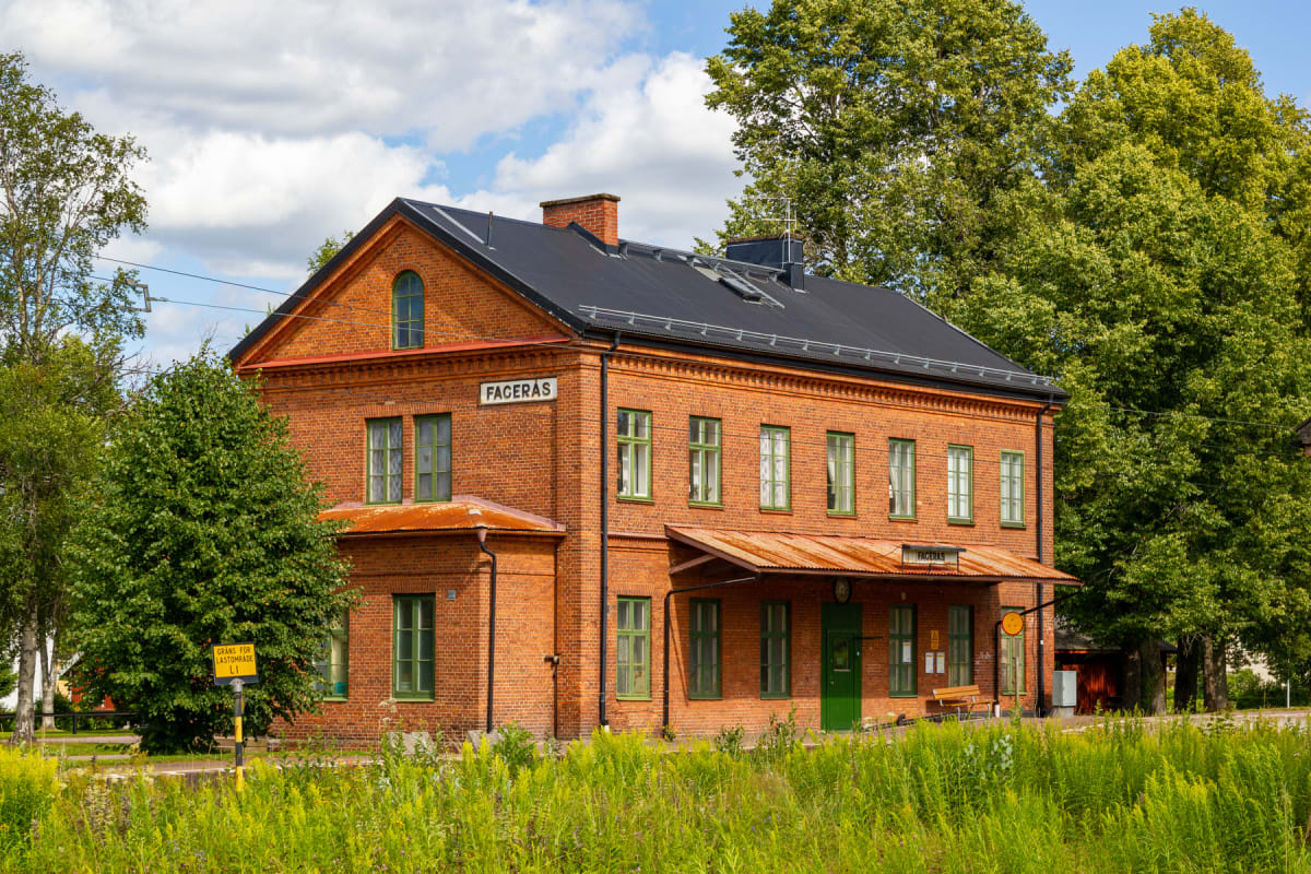 En röd tegelbyggnad, troligen en gammal tågstation, syns framifrån med ett svart tak och grönmålade fönsterkarmar. Namnet "FAGERÅS" står på byggnaden. I förgrunden syns högt grönt gräs och i bakgrunden lummiga träd, allt under en himmel med vita moln